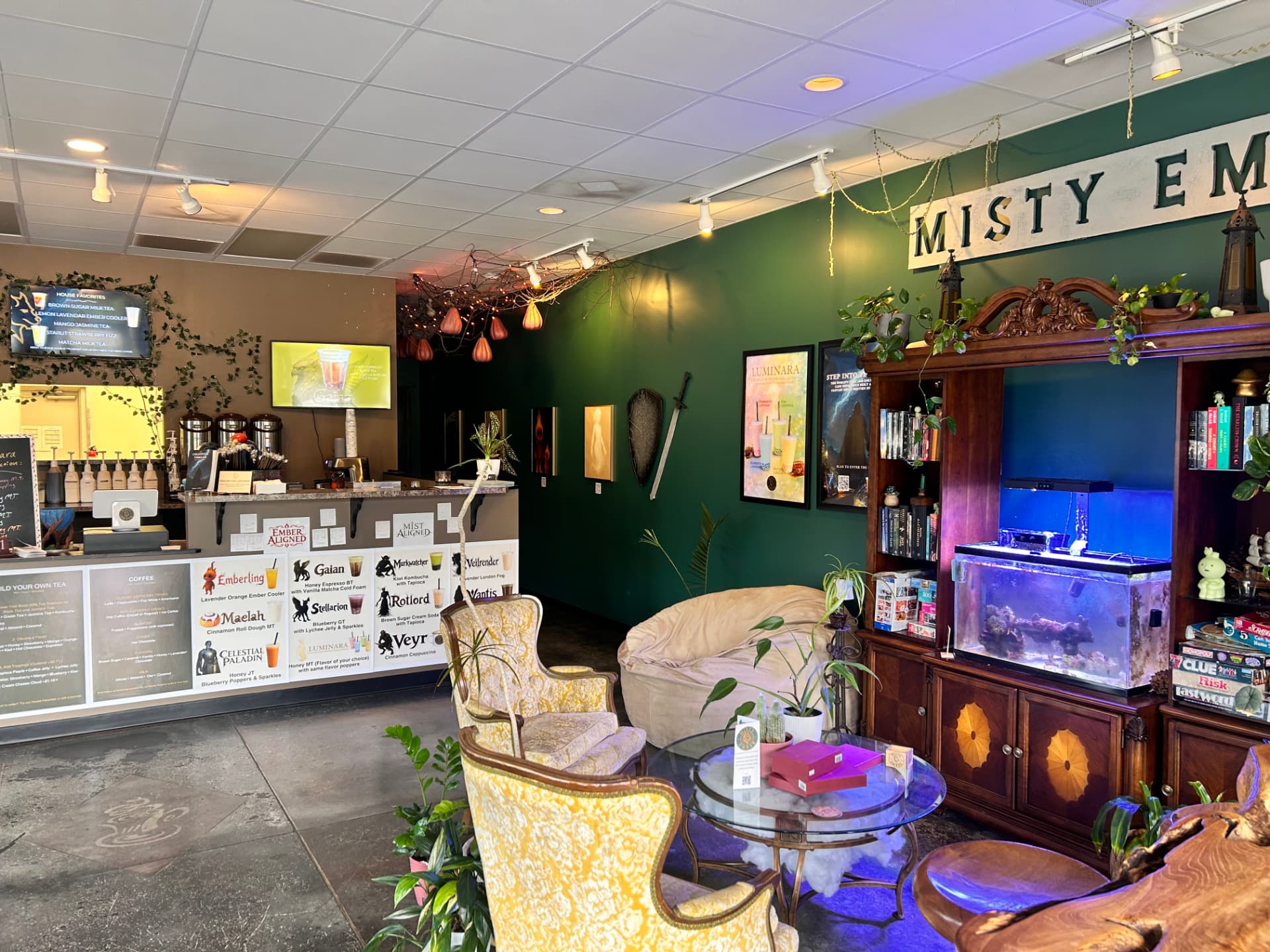 Inside Misty Ember Café: the drink counter with menu boards, yellow armchairs, and a wooden hutch holding books, games, and a lit aquarium