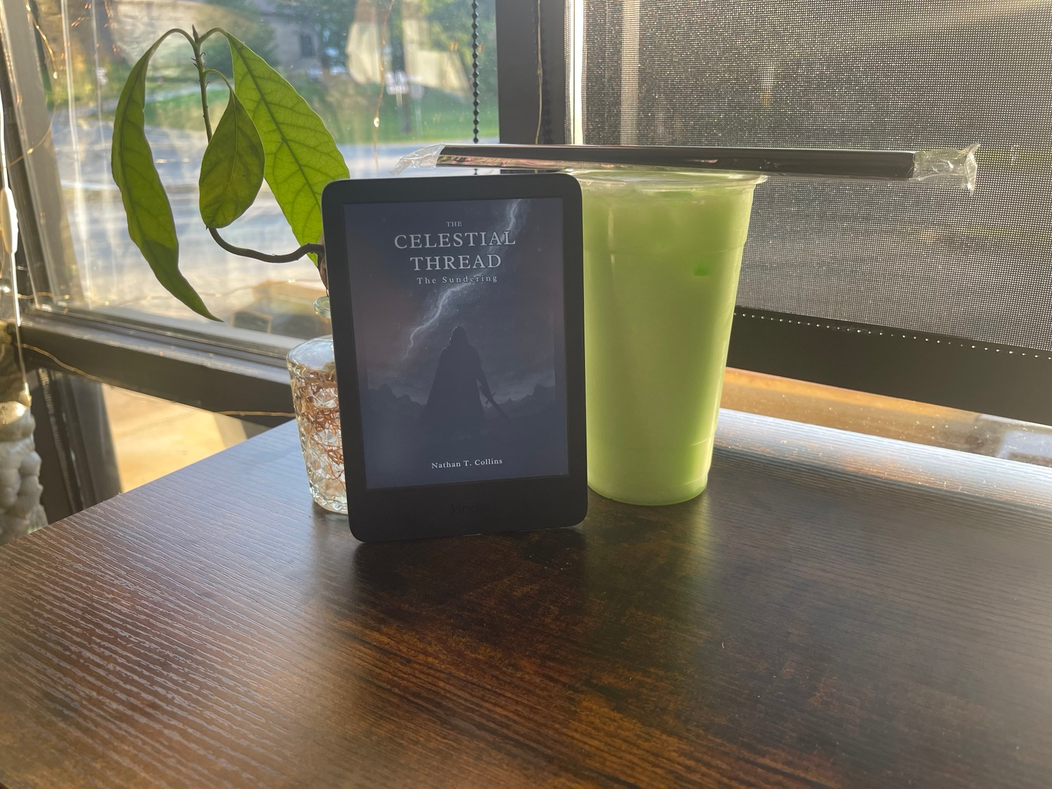 An e-reader showing The Celestial Thread beside an iced green drink on a café table by the window